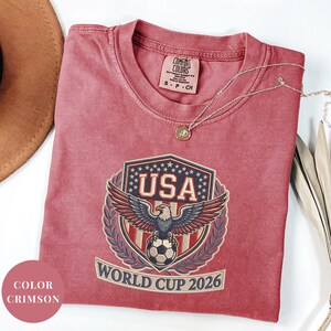 Crimson Comfort Colors 1717 t-shirt featuring a detailed faux embroidery USA World Cup 2026 emblem. The crest shows a bald eagle perched on a soccer ball, surrounded by a laurel wreath and stars. Premium patriotic soccer fan apparel for World Cup 2026.