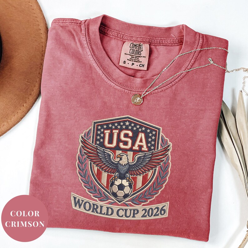 Crimson Comfort Colors 1717 t-shirt featuring a detailed faux embroidery USA World Cup 2026 emblem. The crest shows a bald eagle perched on a soccer ball, surrounded by a laurel wreath and stars. Premium patriotic soccer fan apparel for World Cup 2026.