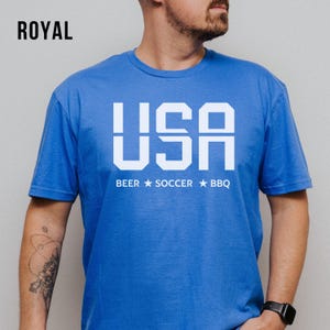 Royal USA T-shirt for Soccer Fans featuring bold block letters and ‘Beer Soccer BBQ’ slogan, patriotic graphic tee for World Cup 2026 and game day wear.