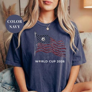 Navy Comfort Colors 1717 t-shirt featuring a vintage-style waving American flag. The design includes a soccer ball integrated into the stars and "WORLD CUP 2026" bold text. High-quality patriotic sports apparel for United States soccer fans and tournament supporters.