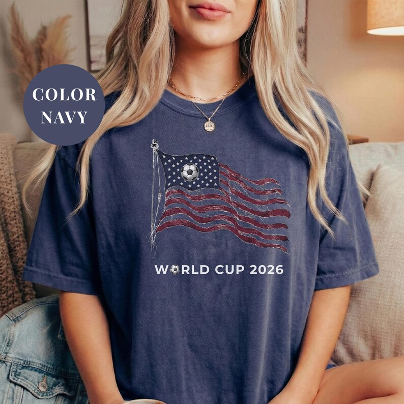 Navy Comfort Colors 1717 t-shirt featuring a vintage-style waving American flag. The design includes a soccer ball integrated into the stars and "WORLD CUP 2026" bold text. High-quality patriotic sports apparel for United States soccer fans and tournament supporters.