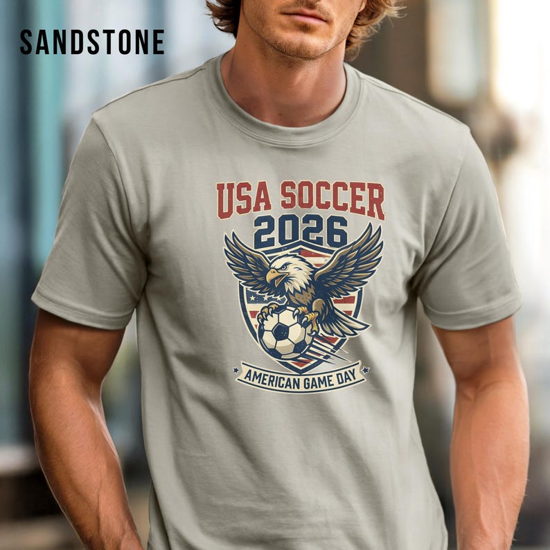 May include: Sandstone-colored t-shirt featuring a graphic of an eagle clutching a soccer ball, with the text "USA SOCCER 2026" and "AMERICAN GAME DAY". The design incorporates red, white, and blue colors, and the shirt has short sleeves.