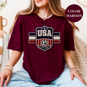 A woman wearing a maroon t-shirt with a shield-shaped design featuring the text "USA" and a soccer ball, along with the year "2020".