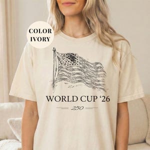 Ivory Comfort Colors 1717 t-shirt featuring a minimalist vintage sketch of a waving American flag. Includes "WORLD CUP '26" and a stylized "250" text celebrating the USA Semiquincentennial. Premium patriotic sports apparel for United States soccer fans and 2026 host nation supporters.