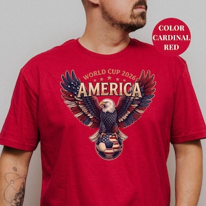 Red t-shirt featuring a patriotic World Cup 2026 America design. A majestic bald eagle with red, white, and blue wings grasps a USA flag pattern soccer ball below "WORLD CUP 2026 AMERICA" text. Premium sports apparel for United States soccer fans and tournament supporters.