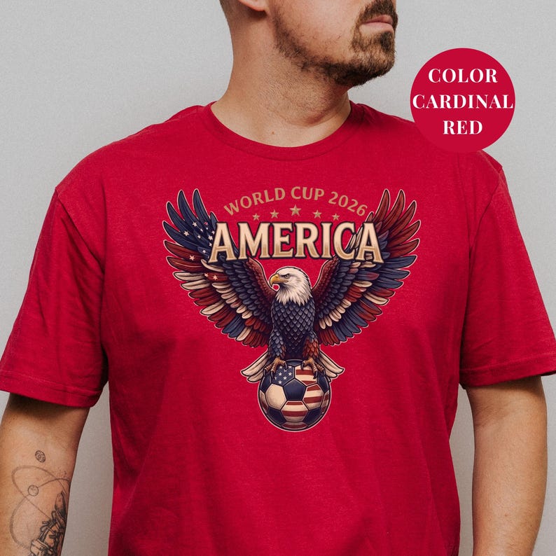 Red t-shirt featuring a patriotic World Cup 2026 America design. A majestic bald eagle with red, white, and blue wings grasps a USA flag pattern soccer ball below "WORLD CUP 2026 AMERICA" text. Premium sports apparel for United States soccer fans and tournament supporters.