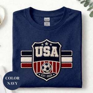 A navy blue t-shirt with a large embroidered shield design featuring the text "USA" and a soccer ball, along with a flag pattern.
