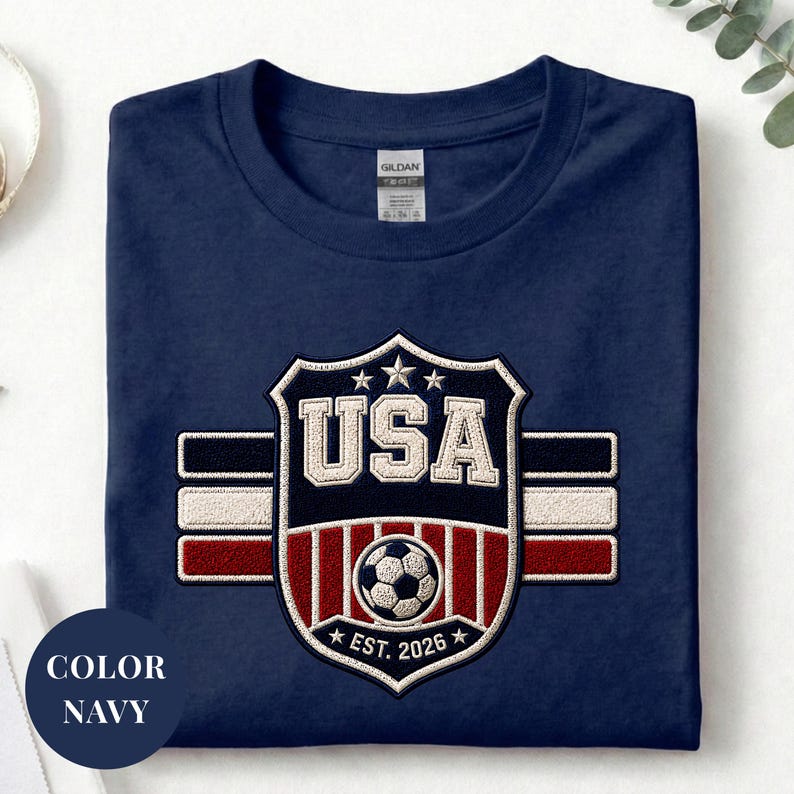 A navy blue t-shirt with a large embroidered shield design featuring the text "USA" and a soccer ball, along with a flag pattern.