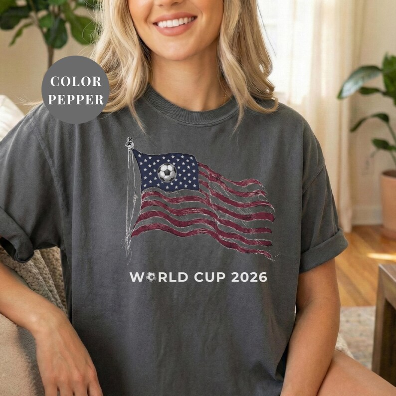 Pepper Comfort Colors 1717 t-shirt featuring a vintage-style waving American flag. The design includes a soccer ball integrated into the stars and "WORLD CUP 2026" bold text. High-quality patriotic sports apparel for United States soccer fans and tournament supporters.
