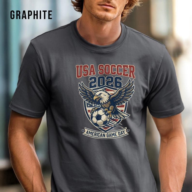 May include: Graphite gray t-shirt with a graphic featuring an eagle, soccer ball, and text "USA SOCCER 2026 AMERICAN GAME DAY". The design incorporates red, white, and blue colors, with the text in a bold, vintage style.