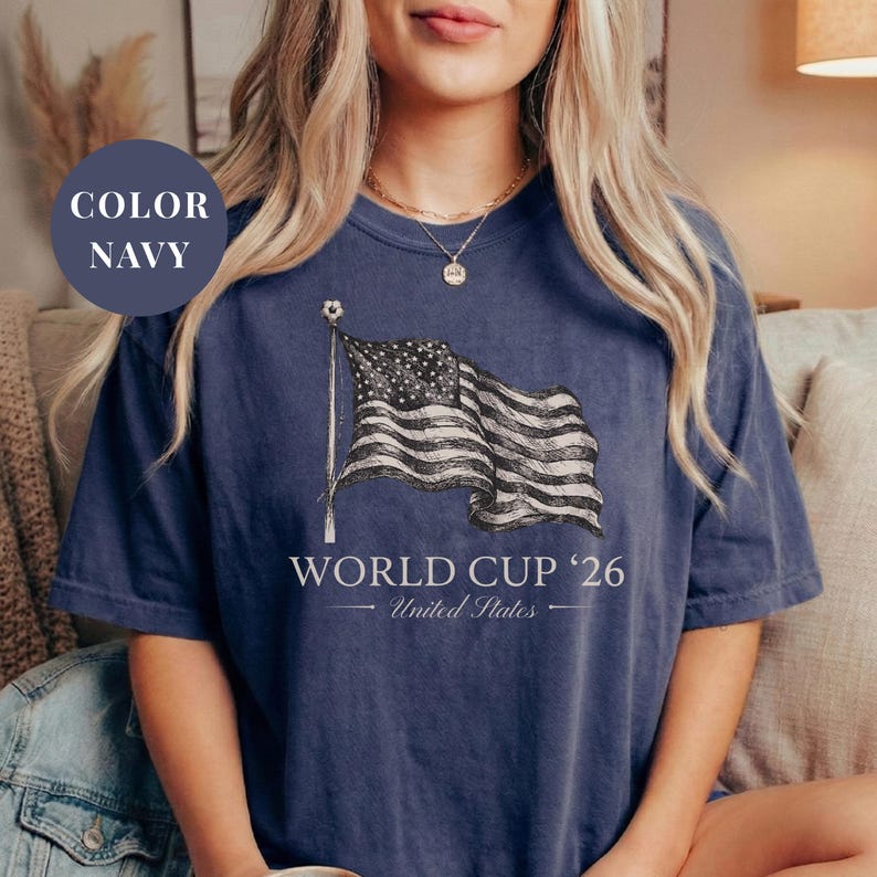 Navy Comfort Colors 1717 t-shirt featuring a vintage hand-drawn sketch of a waving American flag. Includes "WORLD CUP '26" and "United States" text, with a subtle soccer ball detail on the flagpole. High-quality patriotic soccer apparel for the 2026 world tournament fans.