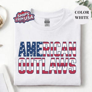 White t-shirt featuring a distressed retro design for "AMERICAN OUTLAWS". The bold collegiate block lettering is filled with a patriotic US national flag pattern of stars and stripes. High-quality vintage-style soccer apparel for passionate United States football supporters and tournament fans.