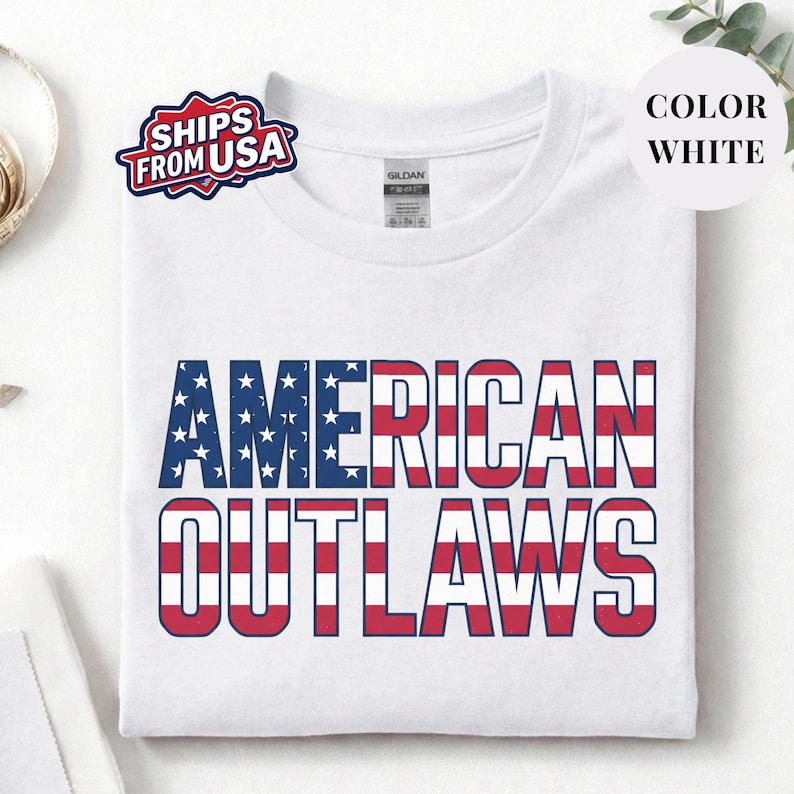 White t-shirt featuring a distressed retro design for "AMERICAN OUTLAWS". The bold collegiate block lettering is filled with a patriotic US national flag pattern of stars and stripes. High-quality vintage-style soccer apparel for passionate United States football supporters and tournament fans.