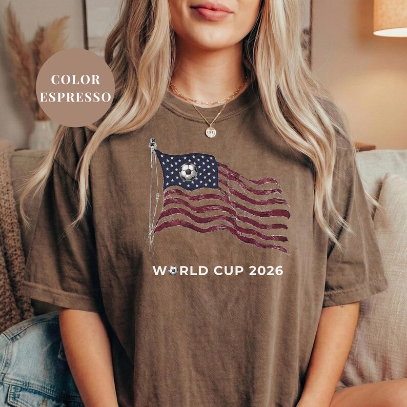 Espresso brown Comfort Colors 1717 t-shirt featuring a vintage-style waving American flag. The design includes a soccer ball integrated into the stars and "WORLD CUP 2026" bold text. High-quality patriotic sports apparel for United States soccer fans and tournament supporters.