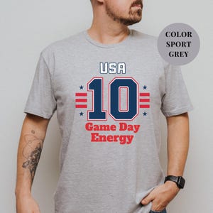 A man wearing a grey t-shirt with a graphic design featuring the text "USA 10 Game Day Energy" and the number 10.
