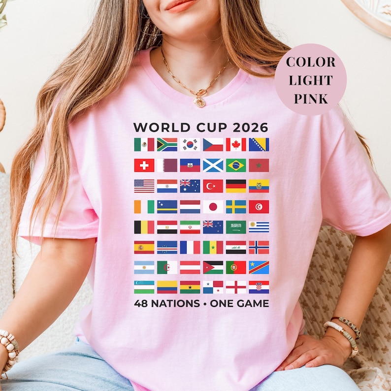 Pink t-shirt featuring a comprehensive grid of 48 national flags for the World Cup 2026. The design includes bold "WORLD CUP 2026" text at the top and the slogan "48 NATIONS • ONE GAME" at the bottom. Premium international soccer fan apparel for the upcoming world tournament in North America.