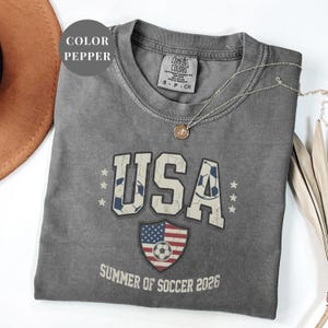 Pepper Comfort Colors 1717 t-shirt with a vintage "USA Summer of Soccer 2026" graphic. Features bold varsity "USA" lettering with soccer ball patterns and a distressed American flag soccer shield. Patriotic apparel for World Cup 2026 fans.