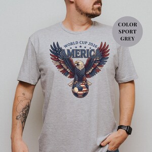 Grey t-shirt featuring a patriotic World Cup 2026 America design. A majestic bald eagle with red, white, and blue wings grasps a USA flag pattern soccer ball below "WORLD CUP 2026 AMERICA" text. Premium sports apparel for United States soccer fans and tournament supporters.