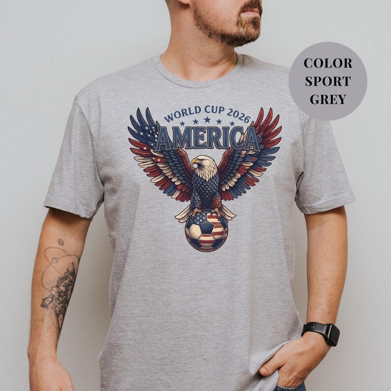 Grey t-shirt featuring a patriotic World Cup 2026 America design. A majestic bald eagle with red, white, and blue wings grasps a USA flag pattern soccer ball below "WORLD CUP 2026 AMERICA" text. Premium sports apparel for United States soccer fans and tournament supporters.