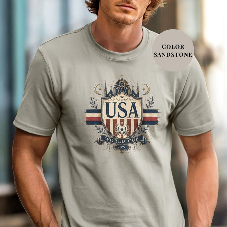 A man wearing a gray t-shirt with a graphic design featuring the text "USA WORLD CUP" and a shield-shaped emblem with a soccer ball and flags.
