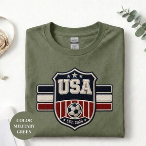 A green military-style t-shirt with a large embroidered patch that says "USA" and features a soccer ball and stripes, along with the text "COLOR MILITARY GREEN" and "EST. 2026" below it.