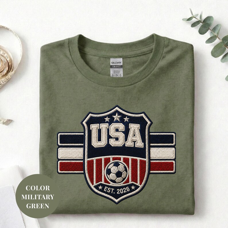 A green military-style t-shirt with a large embroidered patch that says "USA" and features a soccer ball and stripes, along with the text "COLOR MILITARY GREEN" and "EST. 2026" below it.