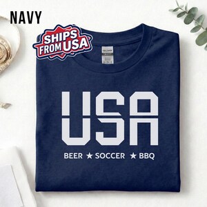 Navy USA T-shirt for Soccer Fans featuring bold block letters and ‘Beer Soccer BBQ’ slogan, patriotic graphic tee for World Cup 2026 and game day wear.
