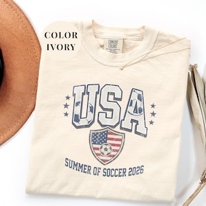 Ivory Comfort Colors 1717 t-shirt with a vintage "USA Summer of Soccer 2026" graphic. Features bold varsity "USA" lettering with soccer ball patterns and a distressed American flag soccer shield. Patriotic apparel for World Cup 2026 fans.