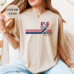 A woman wearing a beige t-shirt with a graphic design featuring a soccer ball and the text "US SOCCER World Cup 26".