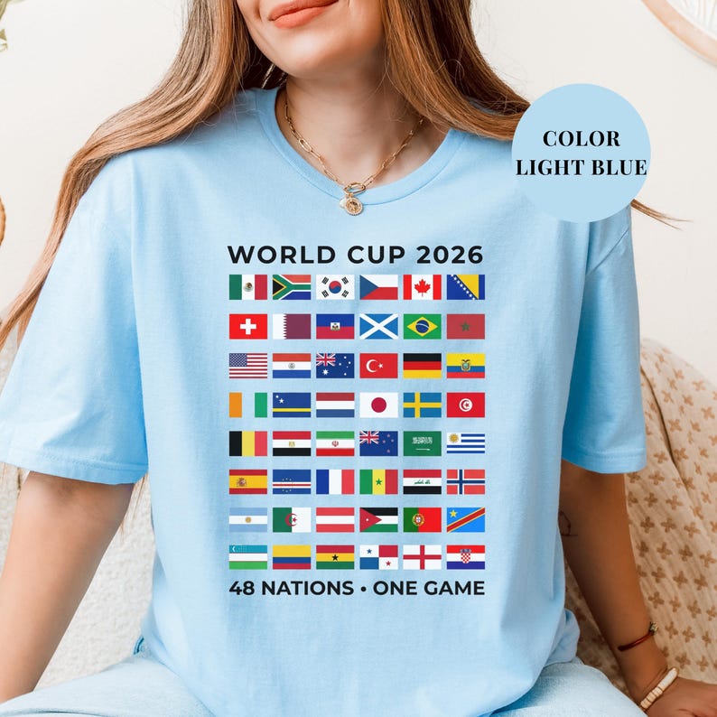 Light Blue t-shirt featuring a comprehensive grid of 48 national flags for the World Cup 2026. The design includes bold "WORLD CUP 2026" text at the top and the slogan "48 NATIONS • ONE GAME" at the bottom. Premium international soccer fan apparel for the upcoming world tournament in North America.