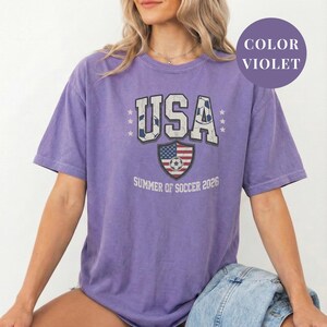 Violet Comfort Colors 1717 t-shirt with a vintage "USA Summer of Soccer 2026" graphic. Features bold varsity "USA" lettering with soccer ball patterns and a distressed American flag soccer shield. Patriotic apparel for World Cup 2026 fans.