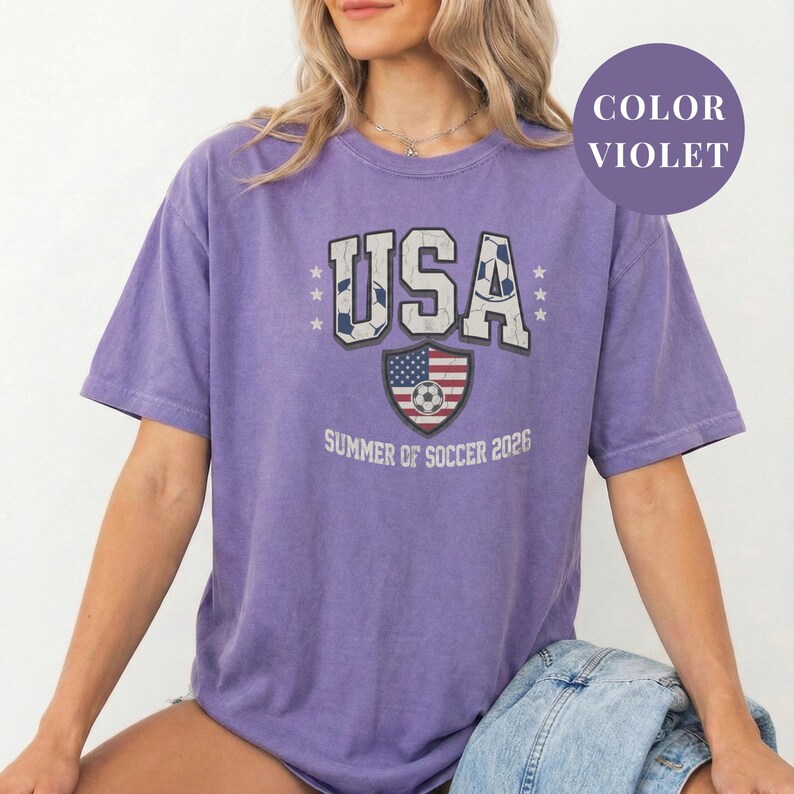 Violet Comfort Colors 1717 t-shirt with a vintage "USA Summer of Soccer 2026" graphic. Features bold varsity "USA" lettering with soccer ball patterns and a distressed American flag soccer shield. Patriotic apparel for World Cup 2026 fans.