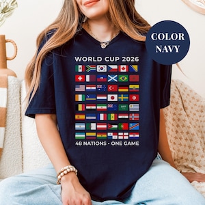 Navy t-shirt featuring a comprehensive grid of 48 national flags for the World Cup 2026. The design includes bold "WORLD CUP 2026" text at the top and the slogan "48 NATIONS • ONE GAME" at the bottom. Premium international soccer fan apparel for the upcoming world tournament in North America.