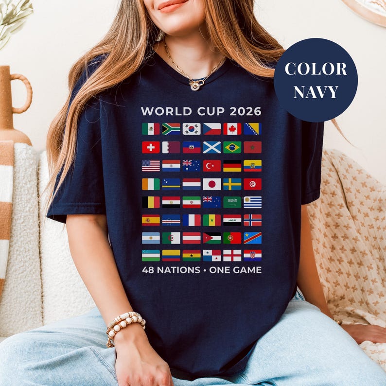 Navy t-shirt featuring a comprehensive grid of 48 national flags for the World Cup 2026. The design includes bold "WORLD CUP 2026" text at the top and the slogan "48 NATIONS • ONE GAME" at the bottom. Premium international soccer fan apparel for the upcoming world tournament in North America.