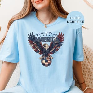 Blue t-shirt featuring a patriotic World Cup 2026 America design. A majestic bald eagle with red, white, and blue wings grasps a USA flag pattern soccer ball below "WORLD CUP 2026 AMERICA" text. Premium sports apparel for United States soccer fans and tournament supporters.