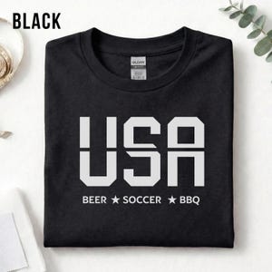 Black USA T-shirt for Soccer Fans featuring bold block letters and ‘Beer Soccer BBQ’ slogan, patriotic graphic tee for World Cup 2026 and game day wear.