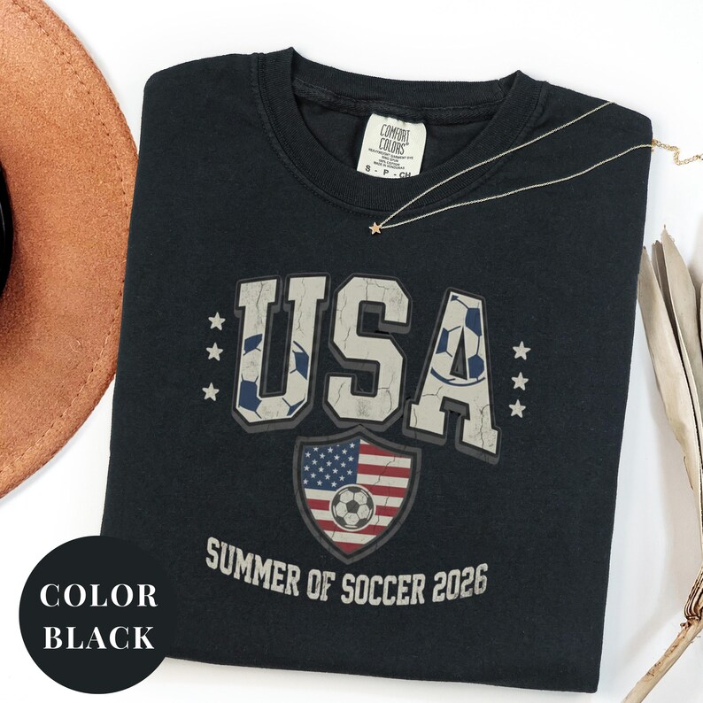 May include: Black t-shirt with a distressed "USA" graphic featuring soccer ball details and stars. The shirt includes a shield with the American flag and a soccer ball, with "Summer of Soccer 2026" text. A gold necklace with a star pendant is also visible.