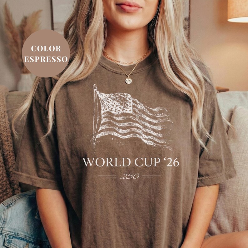 May include: Espresso-colored t-shirt featuring a distressed white graphic of the American flag. Below the flag, the text reads "WORLD CUP '26" with the number "250" underneath. The shirt is a relaxed fit.