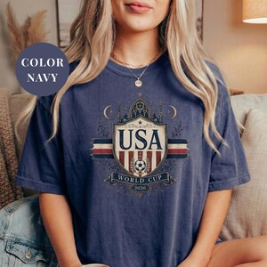 A woman wearing a navy blue t-shirt with a graphic design featuring the text "USA WORLD CUP 2020" and a shield-like emblem.