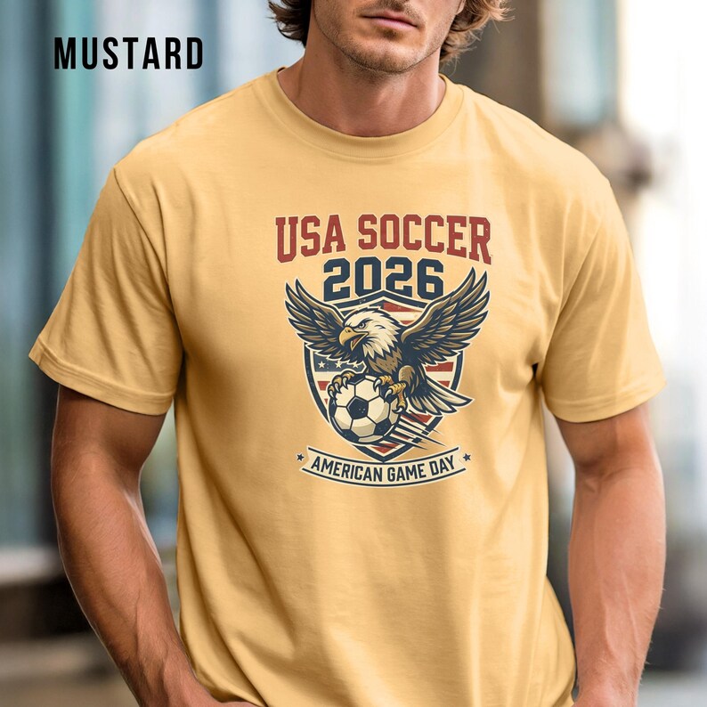 May include: Mustard-colored t-shirt featuring a graphic with an eagle, soccer ball, and the text "USA SOCCER 2026 AMERICAN GAME DAY." The design is in red, white, and blue. The shirt is short-sleeved.