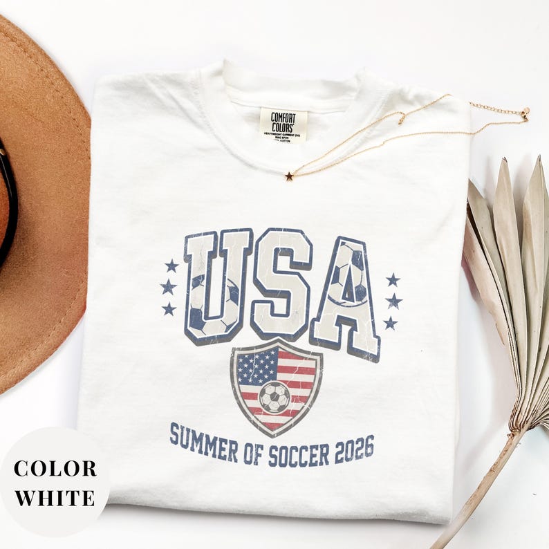White Comfort Colors 1717 t-shirt with a vintage "USA Summer of Soccer 2026" graphic. Features bold varsity "USA" lettering with soccer ball patterns and a distressed American flag soccer shield. Patriotic apparel for World Cup 2026 fans.