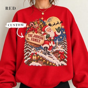Puede incluir: Una sudadera roja de cuello redondo con un estampado gráfico de Santa Claus cargando un saco con la etiqueta THE JOHNSON FAMILY. El diseño incluye un pueblo nevado, una luna llena con la silueta de un trineo y cielos estrellados. Texto RED y CUSTOM.