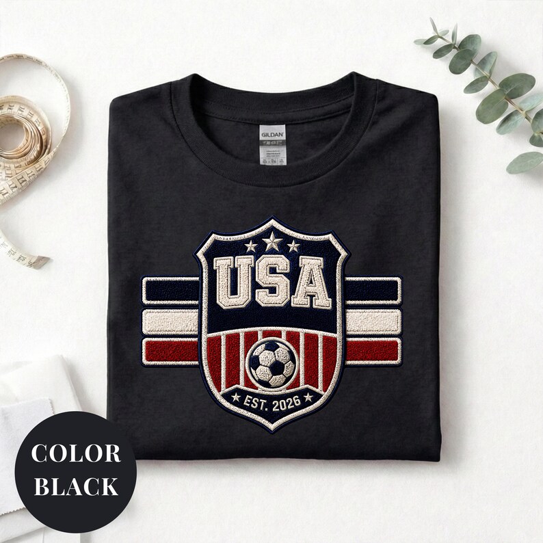 A black t-shirt with a large embroidered patch that says "USA" and features a soccer ball and stripes, along with the text "EST. 2026".