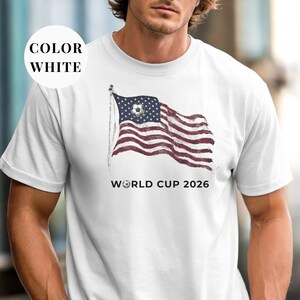 White Comfort Colors 1717 t-shirt featuring a vintage-style waving American flag. The design includes a soccer ball integrated into the stars and "WORLD CUP 2026" bold text. High-quality patriotic sports apparel for United States soccer fans and tournament supporters.