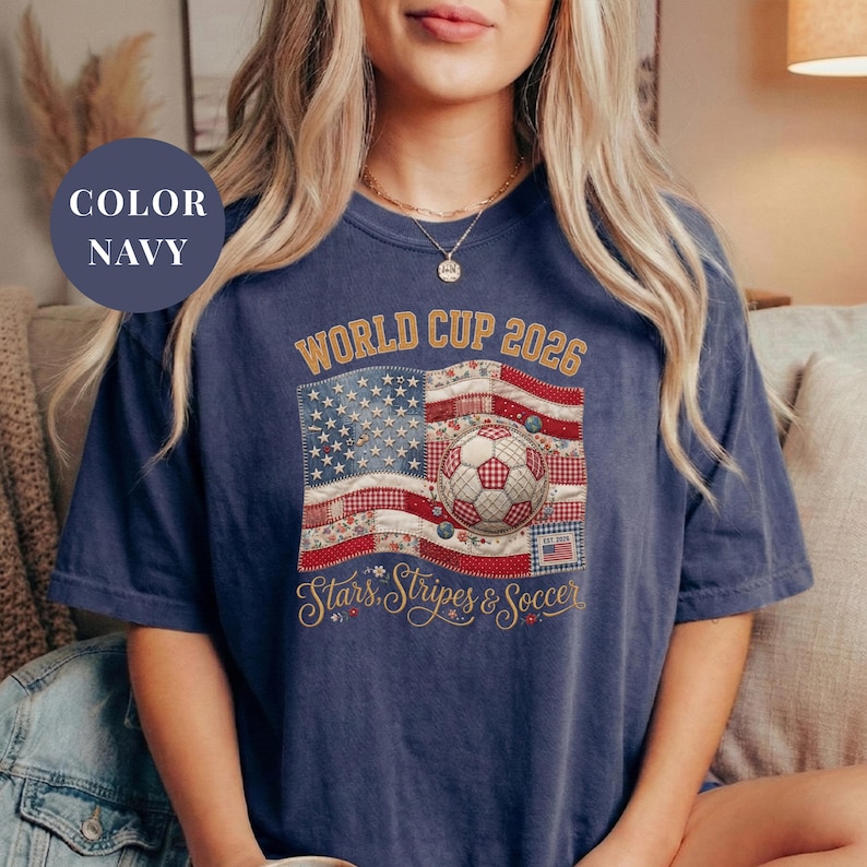 Navy colored t-shirt featuring a vintage patchwork American flag design for World Cup 2026. The faux quilted graphic includes floral and gingham fabric textures, a stitched soccer ball, and "Stars, Stripes & Soccer" text. Unique patriotic sports apparel for women and USA soccer fans.