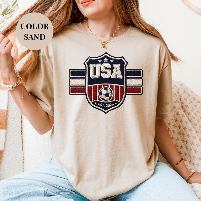 A woman wearing a beige t-shirt with a graphic design of the United States flag and the text "USA" printed on it.