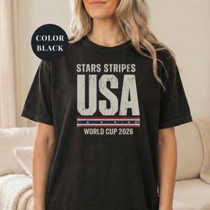 Black t-shirt featuring a distressed vintage "STARS STRIPES USA WORLD CUP 2026" graphic. The design includes weathered block lettering for "USA" and a patriotic red, white, and blue banner with stars. High-quality sports apparel for United States soccer fans and world tournament supporters.