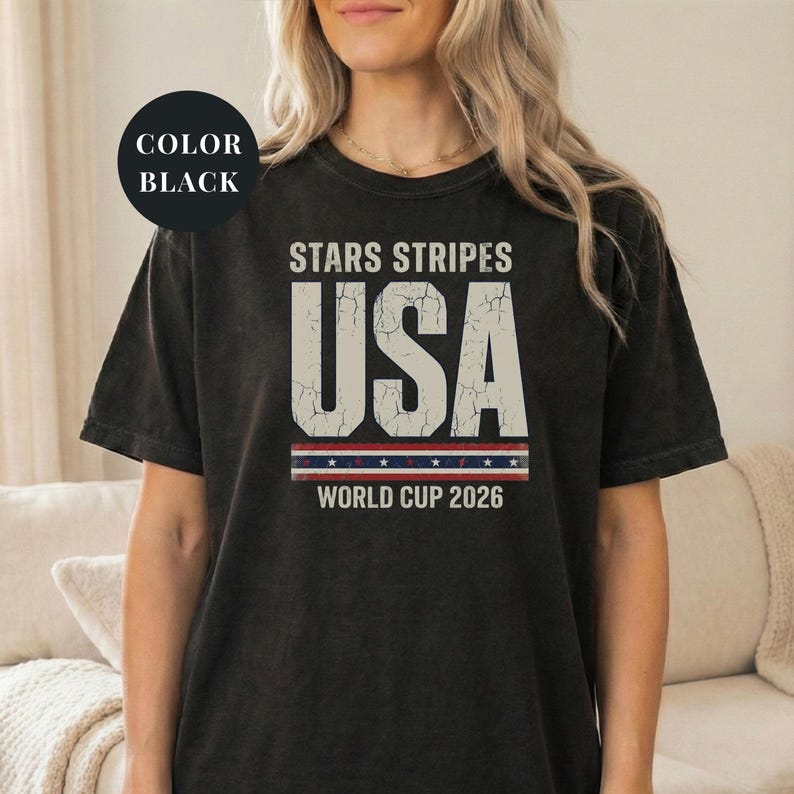 Black t-shirt featuring a distressed vintage "STARS STRIPES USA WORLD CUP 2026" graphic. The design includes weathered block lettering for "USA" and a patriotic red, white, and blue banner with stars. High-quality sports apparel for United States soccer fans and world tournament supporters.