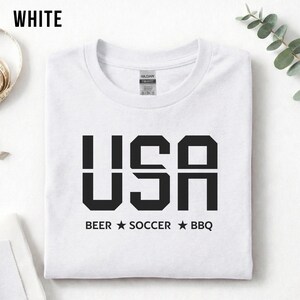 White USA T-shirt for Soccer Fans featuring bold block letters and ‘Beer Soccer BBQ’ slogan, patriotic graphic tee for World Cup 2026 and game day wear.