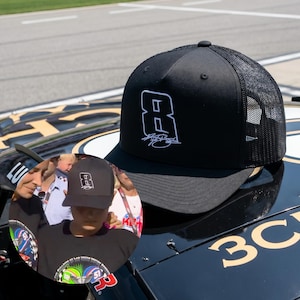 May include: Black trucker hat with a white number 8 and signature logo. The hat has a mesh back and a curved brim. The hat is sitting on a black car.